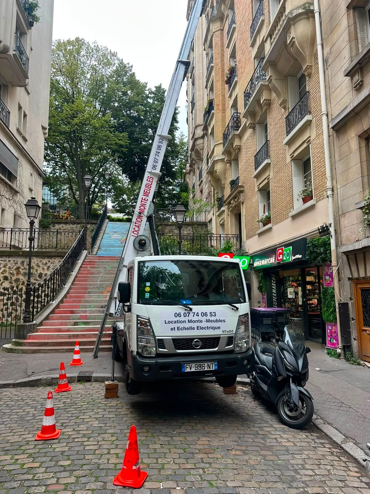 Camion monte-meuble IDEV EXPRESS installé dans une ruelle pavée étroite de Montmartre, bras déployé le long d’un escalier en pente