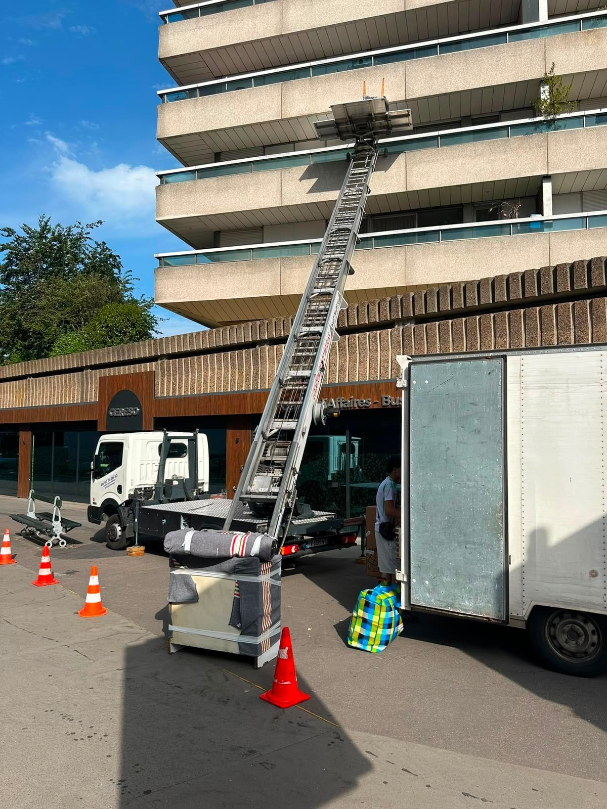 Monte-meuble IDEV EXPRESS déployé depuis son camion vers les étages d’un immeuble, plateau chargé de couvertures de déménagement et cônes de signalisation au sol