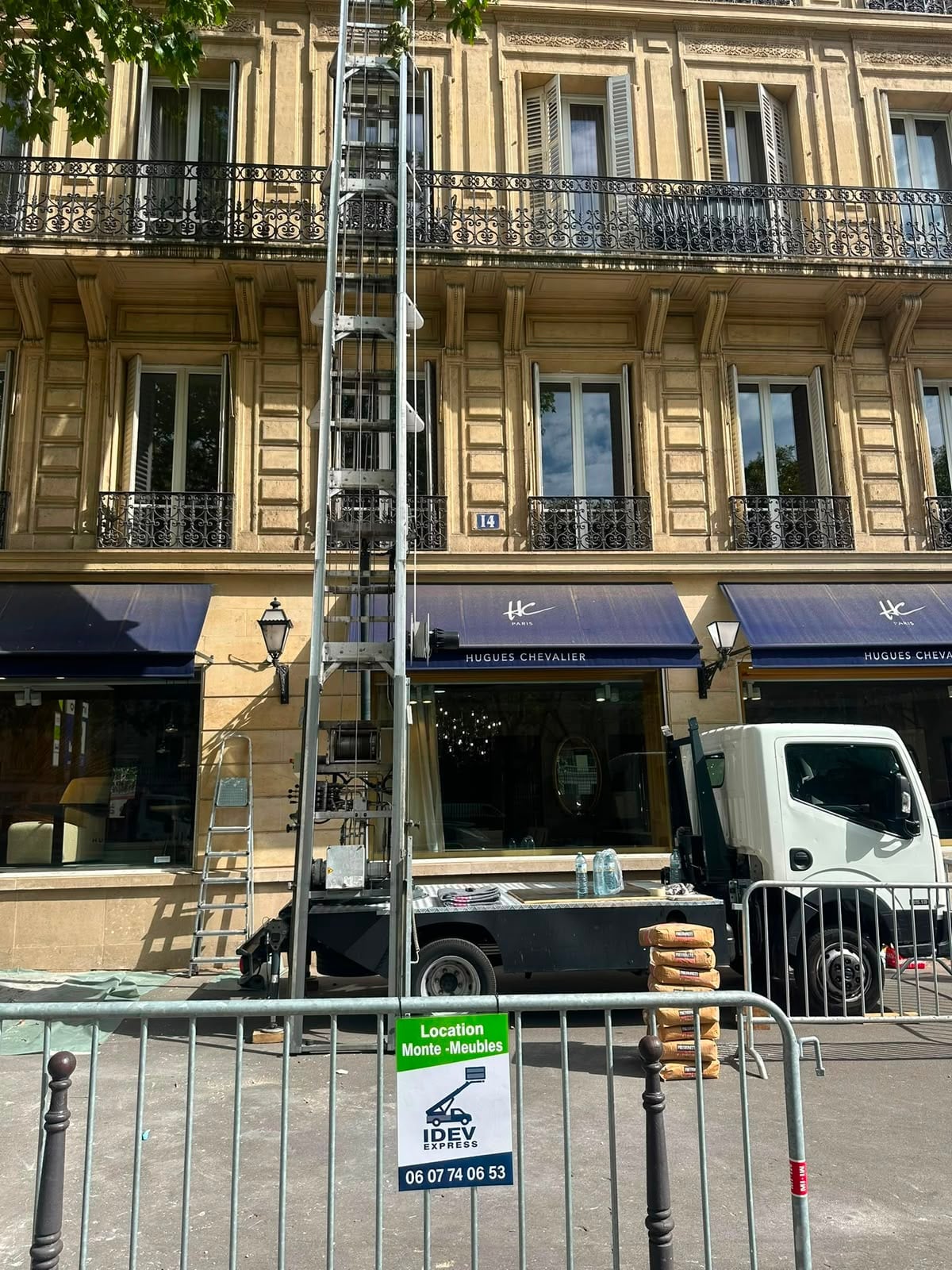 Monte-meuble IDEV EXPRESS dressé le long d’une façade haussmannienne parisienne, sacs de ciment empilés à son pied derrière les barrières de chantier